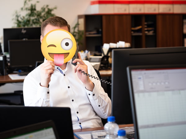 Mann vor Computer am Telefon mit Zunge zeigendem Smiley-Gesicht.