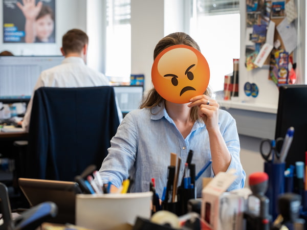 Frau am Arbeitsplatz mit bösem Smiley vor dem Gesicht
